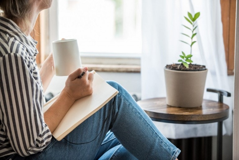 A young woman starts her morning to do something analog like writing in her journal and drinking tea. This is a healthy practice to set a positive mindset for the day.