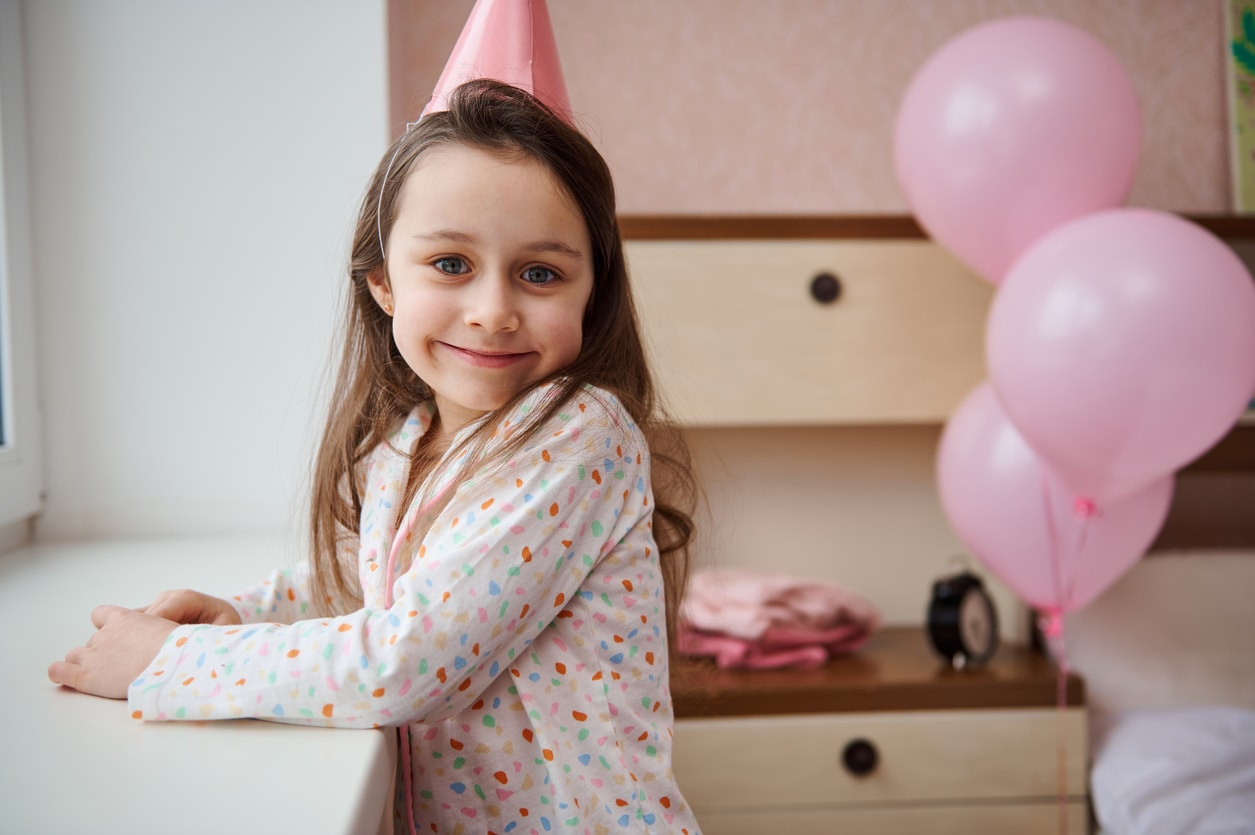 Lovely little child, cute birthday girl in stylish pajamas and pink festive hat, smiles looking into the camera while standing by window in her bedroom decorated with pink inflatable helium balloons