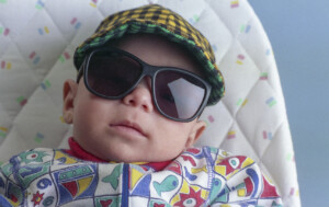 One cute Latino baby boy lying on tabletop baby bouncer wearing a checkered cap and sunglasses.