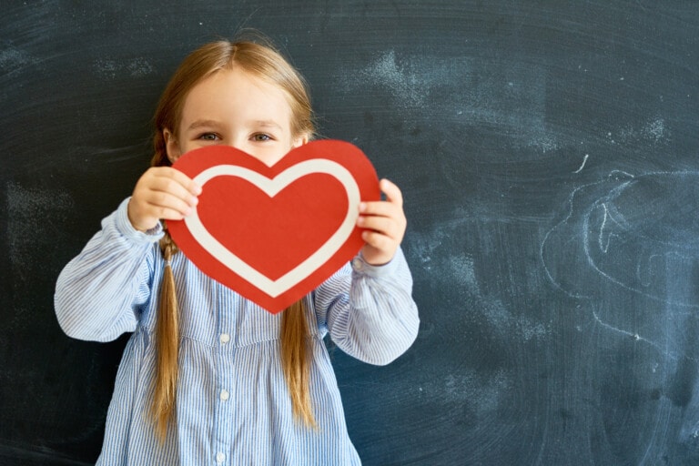 Portrait of adorable little girl hiding her face behind a red paper heart and looking at camera posing against blackboard in school.