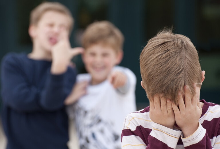 An upset elementary school boy hides his face while being bullied by two other boys in the background. Shot in front of their elementary school.