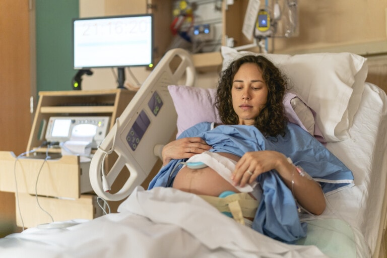 A young ethnic woman is laying in a hospital bed and is having the baby's heartbeat and contractions monitored during the labour and delivery process while she try's to rest. She's wearing a blue hospital gown.