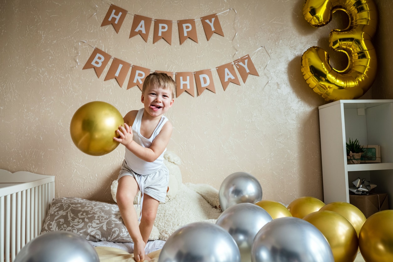 Happy little boy jumping and laughs on bed with balloons in room decorated for birthday celebration. The concept of a birthday.