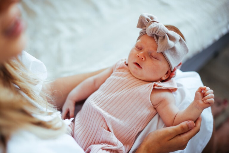Young mother holding her sleeping baby girl in her lap. Baby is wearing a onesie and a bow headband.