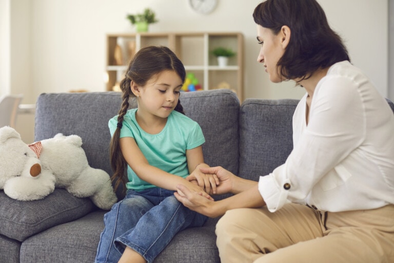 Young mother sitting on sofa with daughter, holding her hands and talking to her seriously at home with room interior at background. Solving problems in children education concept