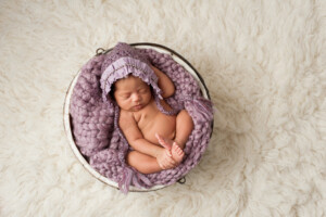 A four week old newborn baby girl sleeping in a little, wooden bucket. She is wearing a lavender colored bonnet. Shot in the studio on a white, flokati rug background.